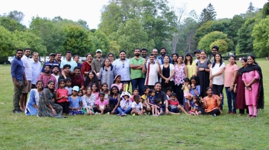 Large community gathering of families and children enjoying fellowship at annual picnic in beautiful green park setting with lush trees